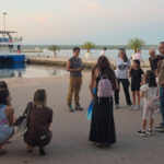 Grupa ljudi, djece i odraslih, okupljena je, na obali, oko jedne osobe koja im govori.